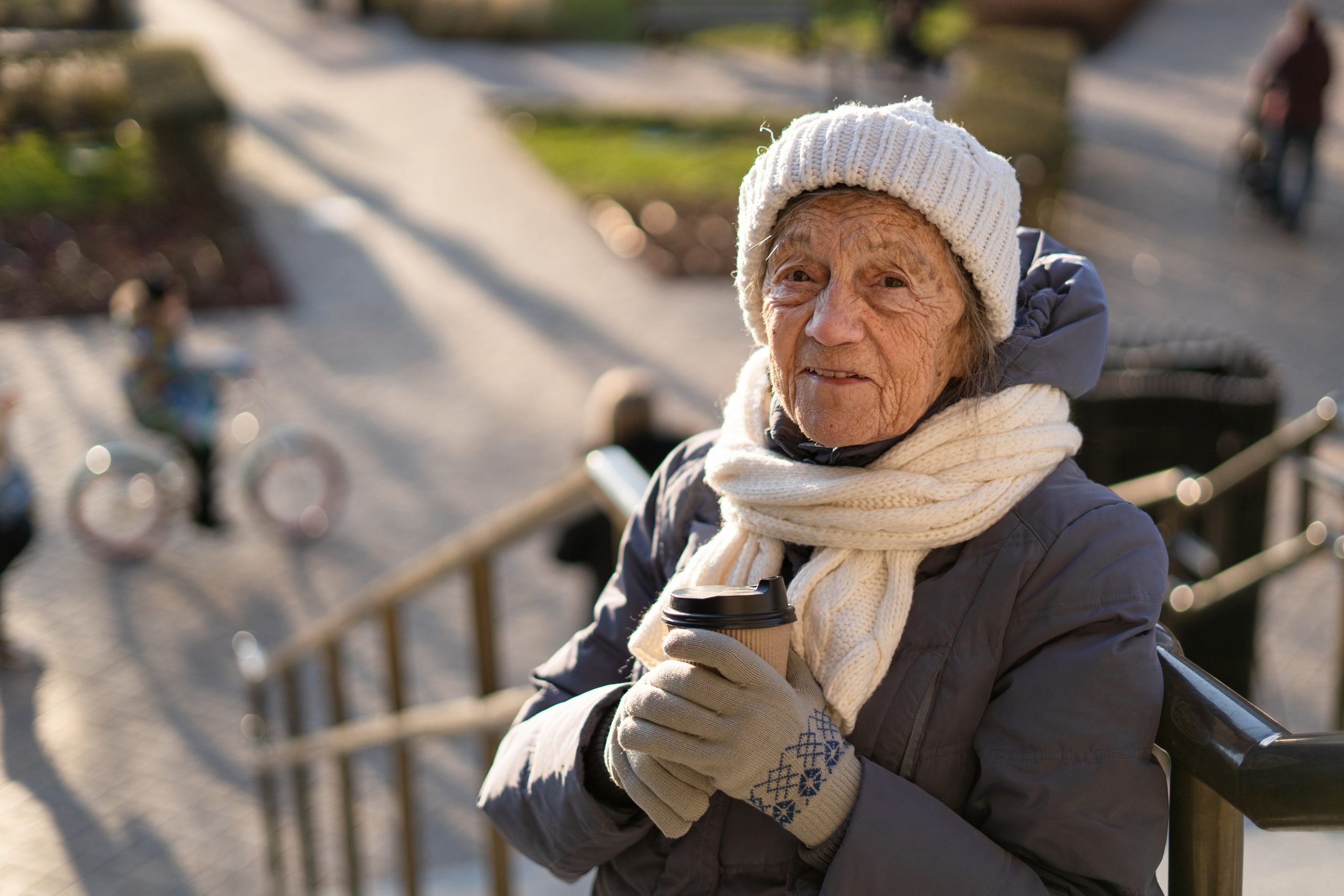 Cute senior woman in city drinks coffee with herself in winter coat and warm hat, scarf and knitted gloves in sunny winter weather. Handsome old female with paper cup of morning coffee to go