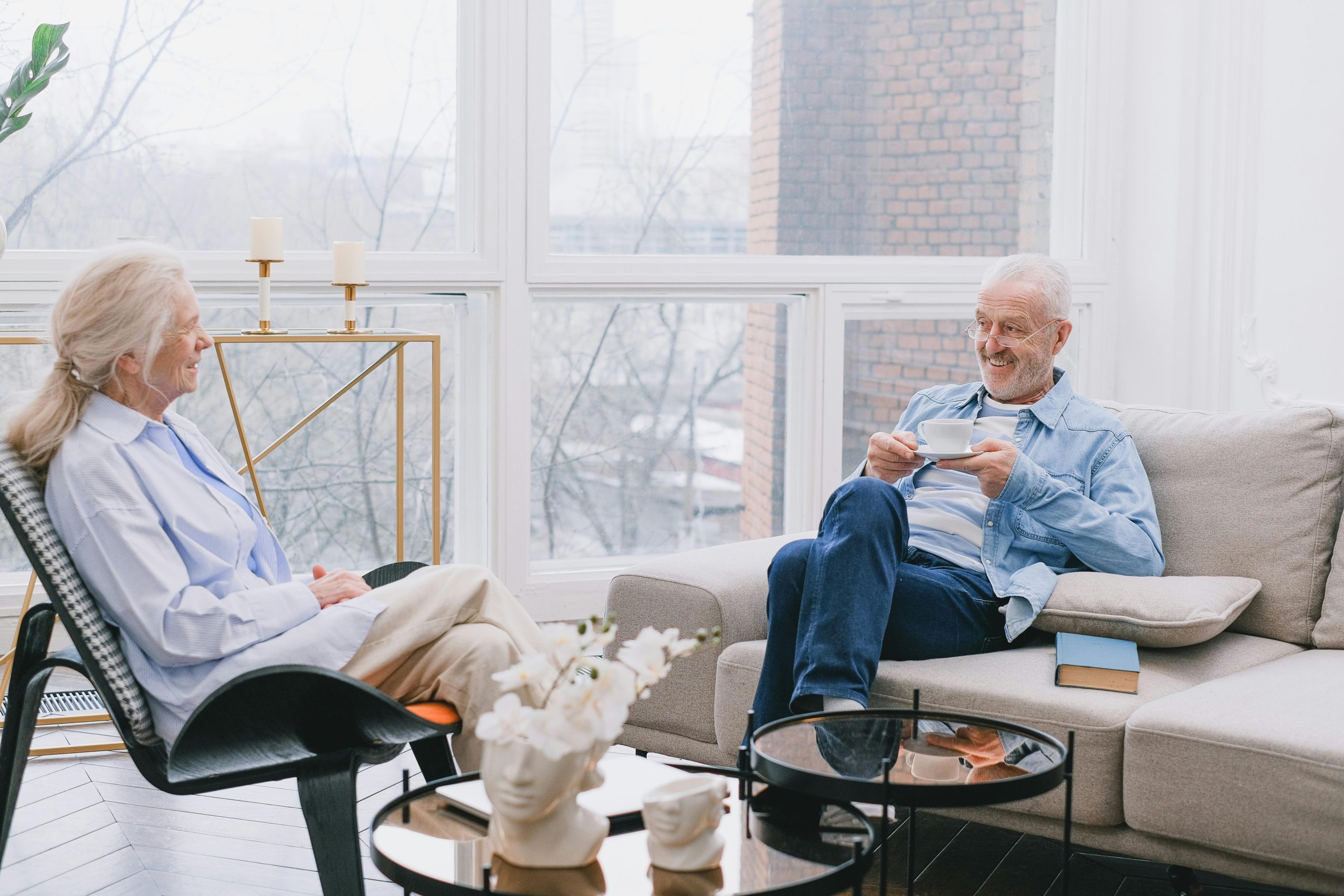 An elderly man and woman sitting together in a bright living room, drinking tea